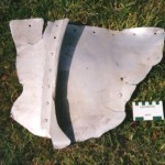 A torn aluminium plate measuring 52cm by 41cm and 3mm thick, riveted to an “L” strut with round headed aluminium rivets. Scale in cm and inches.